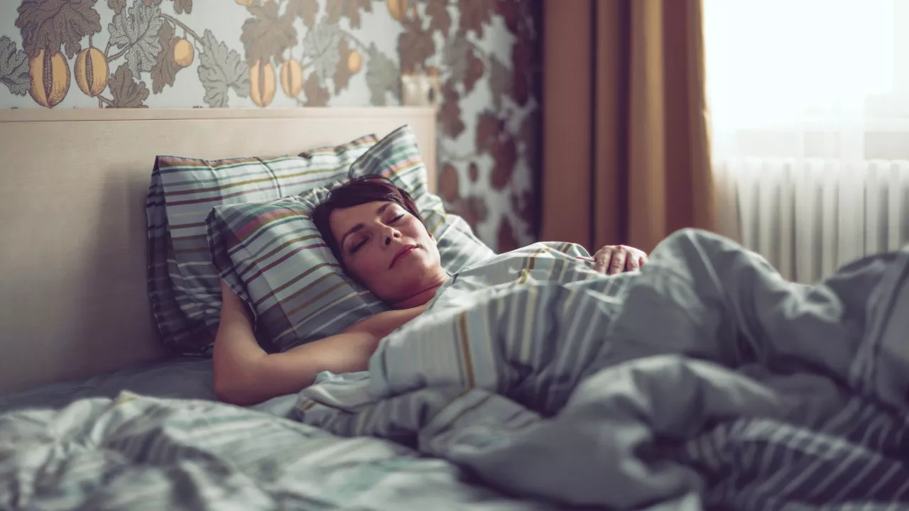 Hiki yöllä saattaa piinava vaihdevuosi-ikäistä. Hymyilevä nainen makaa sängyssä peiton alla.