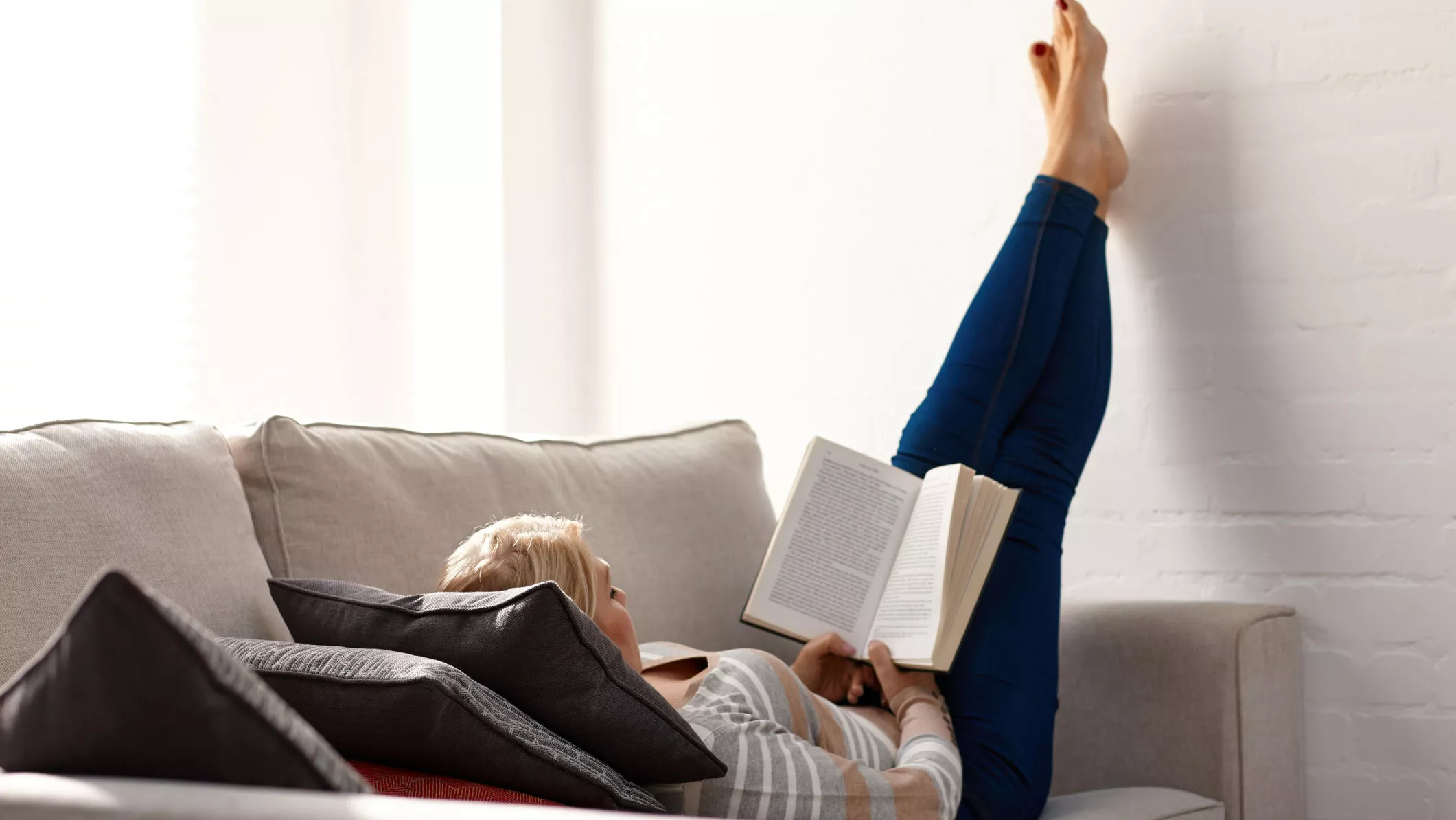 Nainen makaa sohvalla jalat ylhäällä ja lukee kirjaa