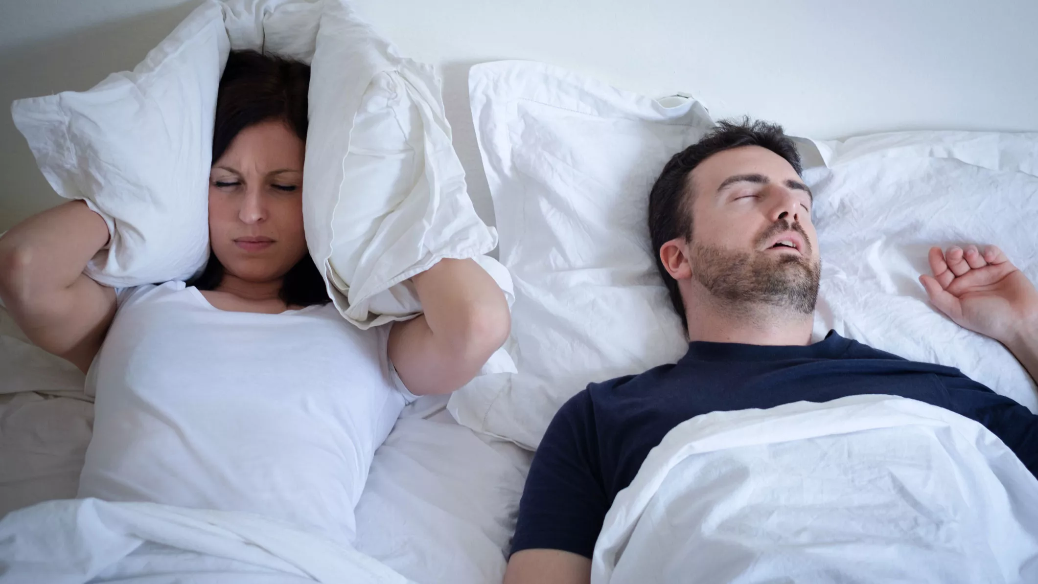 Nainen ja mies nukkumassa, mies kuorsaa, nainen pitää tyynyä korvillaan ja miettii, miten kuorsaaminen saataisiin loppumaan.