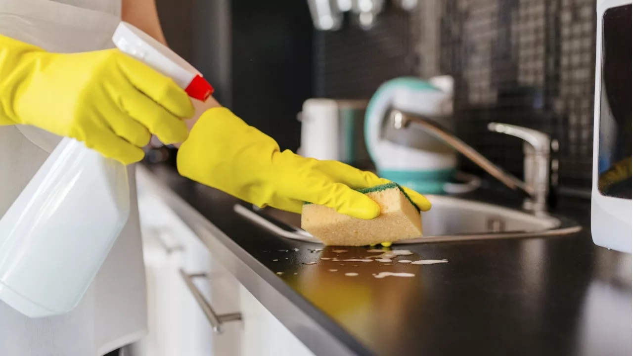 ihminen siivoaa keittiötasoa kumihanskat, suihkepullo ja keittiösieni kädessä.