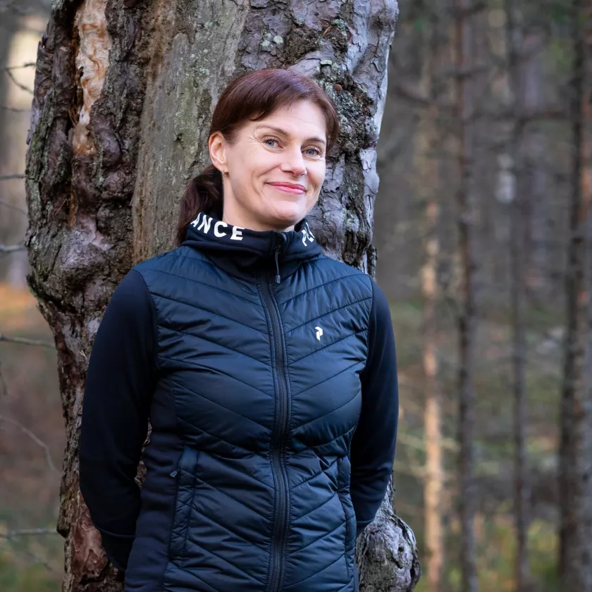 Tiita-Maria nojaa puuhun metsässä ja hymyilee.  