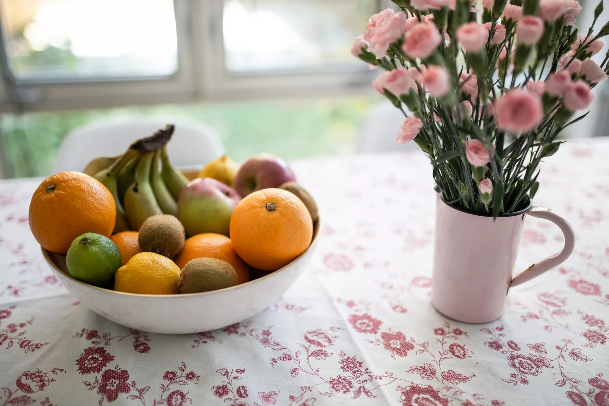Kuvassa on pöydällä pöytäliinan päällä hedelmäkulho (vas.) ja neilikkakimppu (oik.)