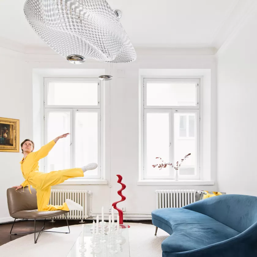 Atte Kilpinen tekee arabesque-tanssiliikkeen sohvatuolilla (vas.) ikkunoiden edessä. Katosta roikkuu Cosmic Angel -valaisin. Oikeassa alakulmassa on sininen sohva.