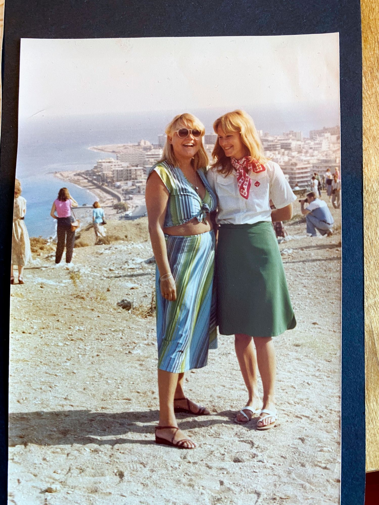 Kaksi iloista naista seisoo kukkulalla, takana näkyy meri ja Rodoksen lomakohde rantoineen.