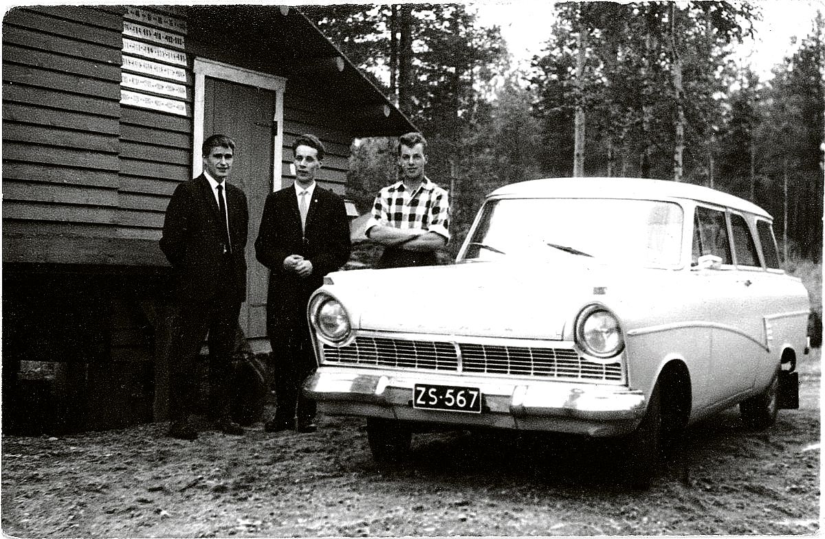 Kolme miestä seisoo valkoisen auton ja maitolavan välissä.